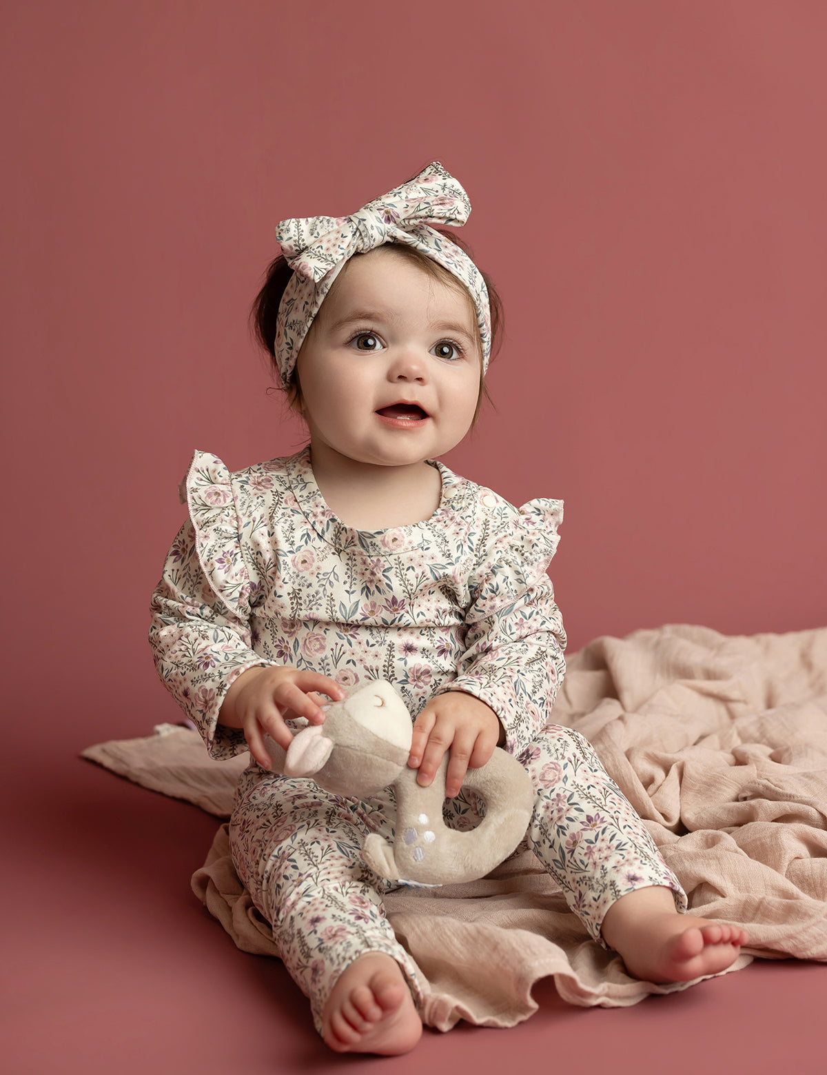 A baby wears the Mabel and Honey Soft Meadow Long Sleeve Floral Ruffle Trim Bodysuit, paired with a matching headband, while sitting on a soft pink blanket and holding a plush toy against a solid dusty pink background—an ideal cozy photo outfit crafted from soft stretch jersey.