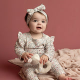 A baby wears the Mabel and Honey Soft Meadow Long Sleeve Floral Ruffle Trim Bodysuit, paired with a matching headband, while sitting on a soft pink blanket and holding a plush toy against a solid dusty pink background—an ideal cozy photo outfit crafted from soft stretch jersey.