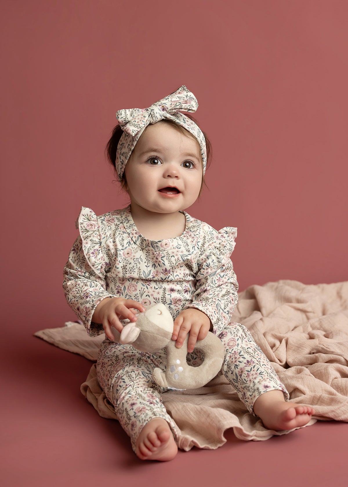 A baby wears the Mabel and Honey Soft Meadow Long Sleeve Floral Ruffle Trim Bodysuit, paired with a matching headband, while sitting on a soft pink blanket and holding a plush toy against a solid dusty pink background—an ideal cozy photo outfit crafted from soft stretch jersey.
