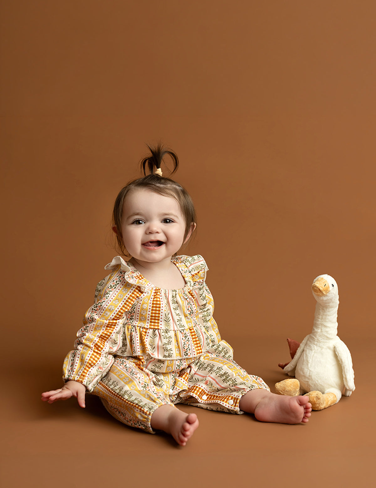 A smiling baby wearing the Mabel and Honey Buttercup Blossom Long Sleeve Striped Floral Ruffle Collar Bubble Romper sits on a brown floor next to a white stuffed duck toy, with a light brown background behind them.