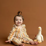 A smiling baby wearing the Mabel and Honey Buttercup Blossom Long Sleeve Striped Floral Ruffle Collar Bubble Romper sits on a brown floor next to a white stuffed duck toy, with a light brown background behind them.