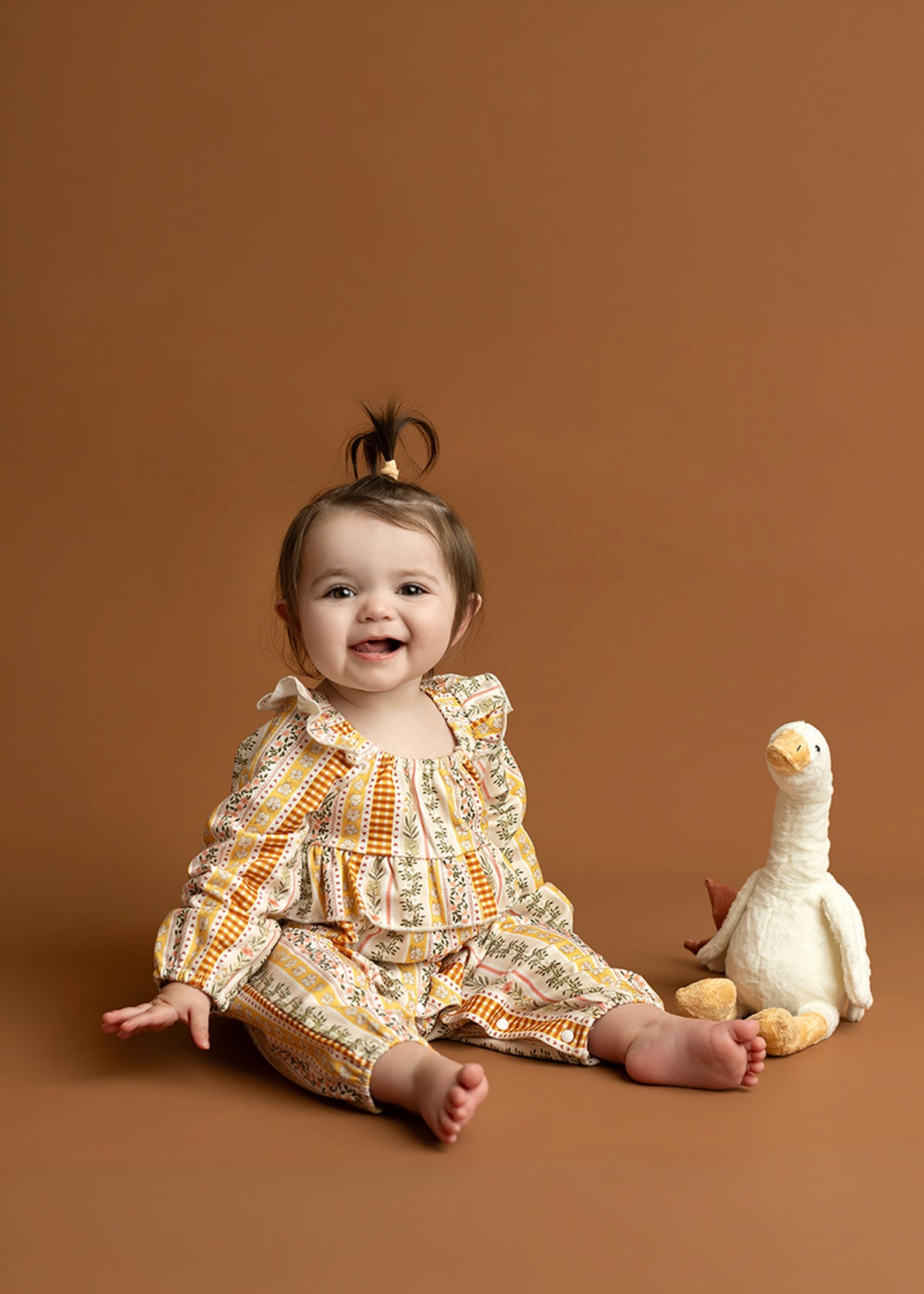 A smiling baby wearing the Mabel and Honey Buttercup Blossom Long Sleeve Striped Floral Ruffle Collar Bubble Romper sits on a brown floor next to a white stuffed duck toy, with a light brown background behind them.