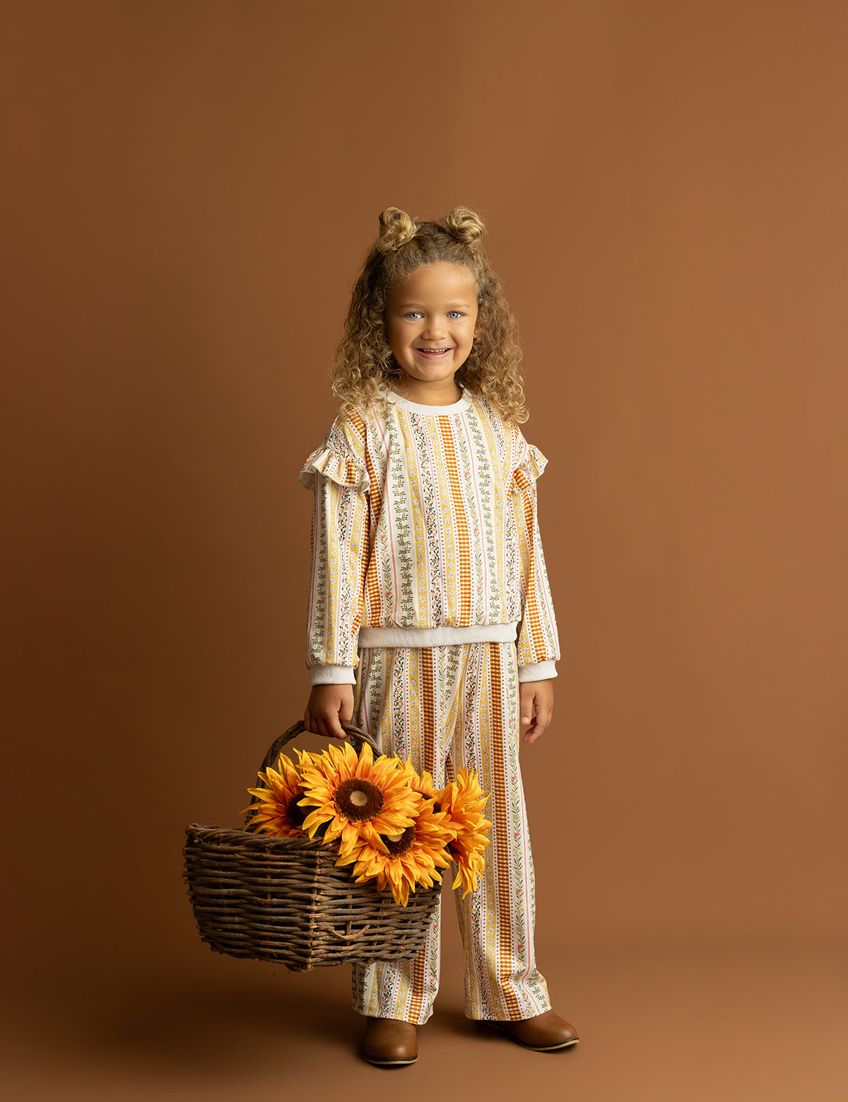 A smiling child with curly hair wears the Mabel and Honey Buttercup Blossom Long Sleeve Striped Ruffle Shoulder Sweater, standing against a brown background in brown shoes and holding a basket of large artificial sunflowers.