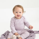 A baby with light brown hair sits on a lavender blanket, wearing Mabel and Honey’s Soft Meadow Solid Ribbed Cuff Joggers in a matching lavender shade. Made from a soft cotton blend with an elastic waistband, the joggers offer both comfort and style as the baby holds a small stuffed toy. The background is plain and white.