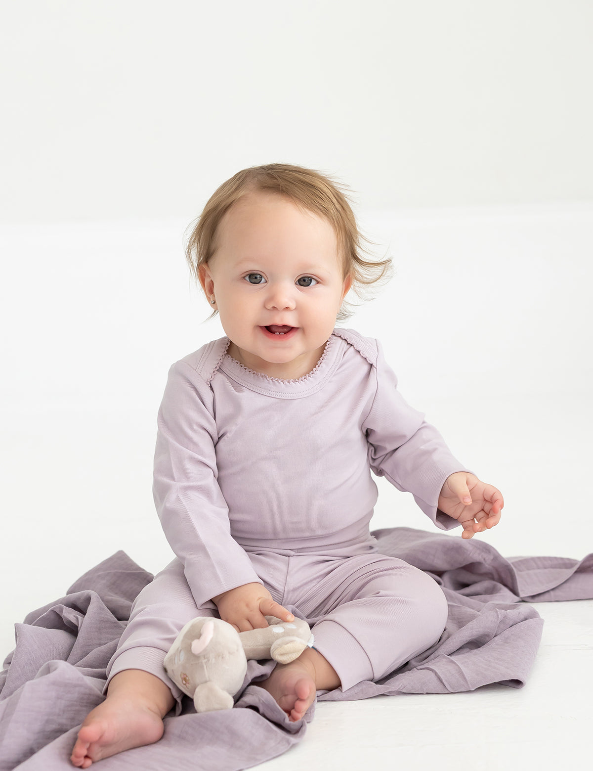 A baby with light brown hair sits on a lavender blanket, wearing Mabel and Honey’s Soft Meadow Solid Ribbed Cuff Joggers in a matching lavender shade. Made from a soft cotton blend with an elastic waistband, the joggers offer both comfort and style as the baby holds a small stuffed toy. The background is plain and white.