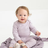 A baby with light brown hair sits on a lavender blanket, wearing Mabel and Honey’s Soft Meadow Solid Ribbed Cuff Joggers in a matching lavender shade. Made from a soft cotton blend with an elastic waistband, the joggers offer both comfort and style as the baby holds a small stuffed toy. The background is plain and white.