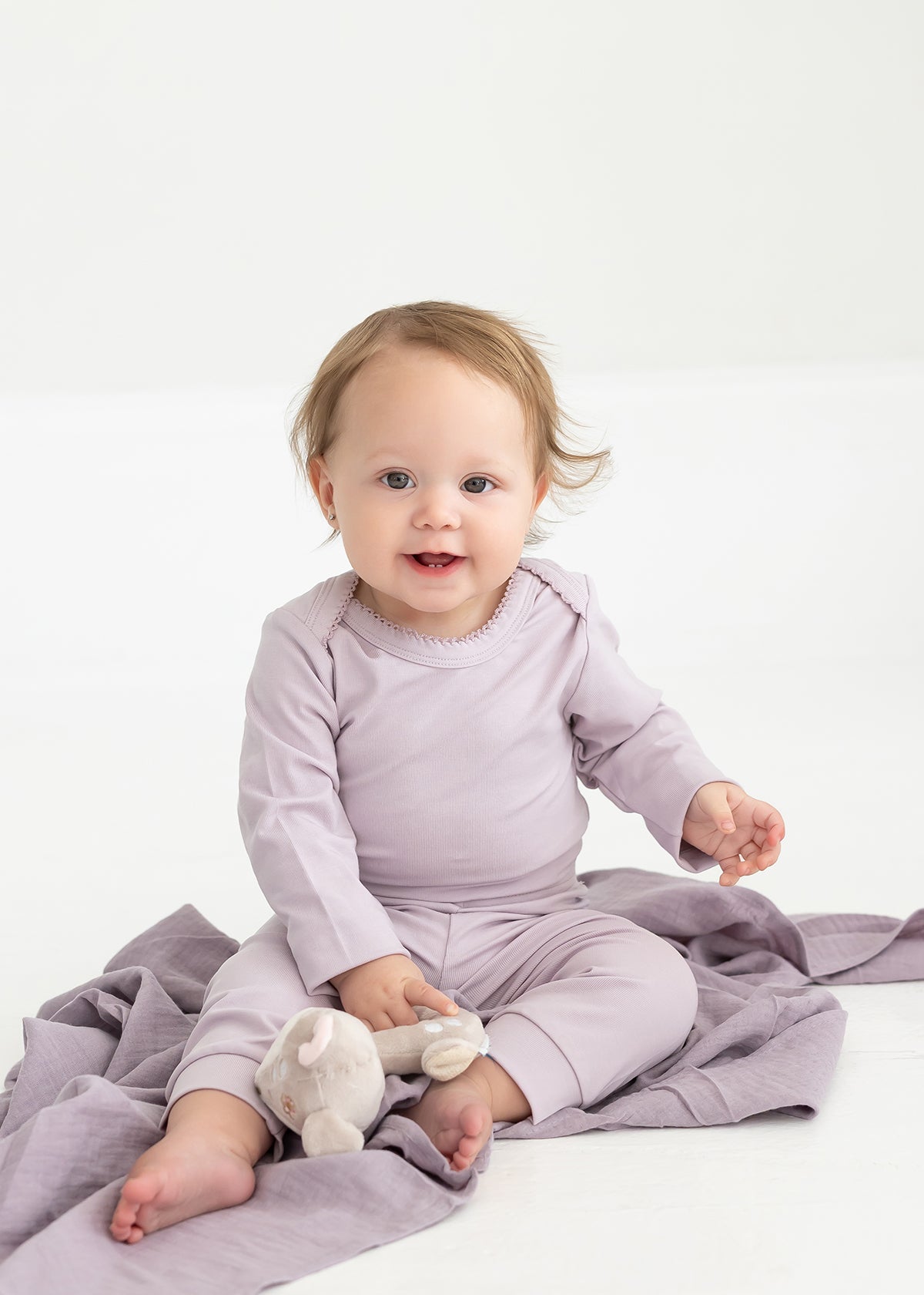 A baby with light brown hair sits on a lavender blanket, wearing Mabel and Honey’s Soft Meadow Solid Ribbed Cuff Joggers in a matching lavender shade. Made from a soft cotton blend with an elastic waistband, the joggers offer both comfort and style as the baby holds a small stuffed toy. The background is plain and white.
