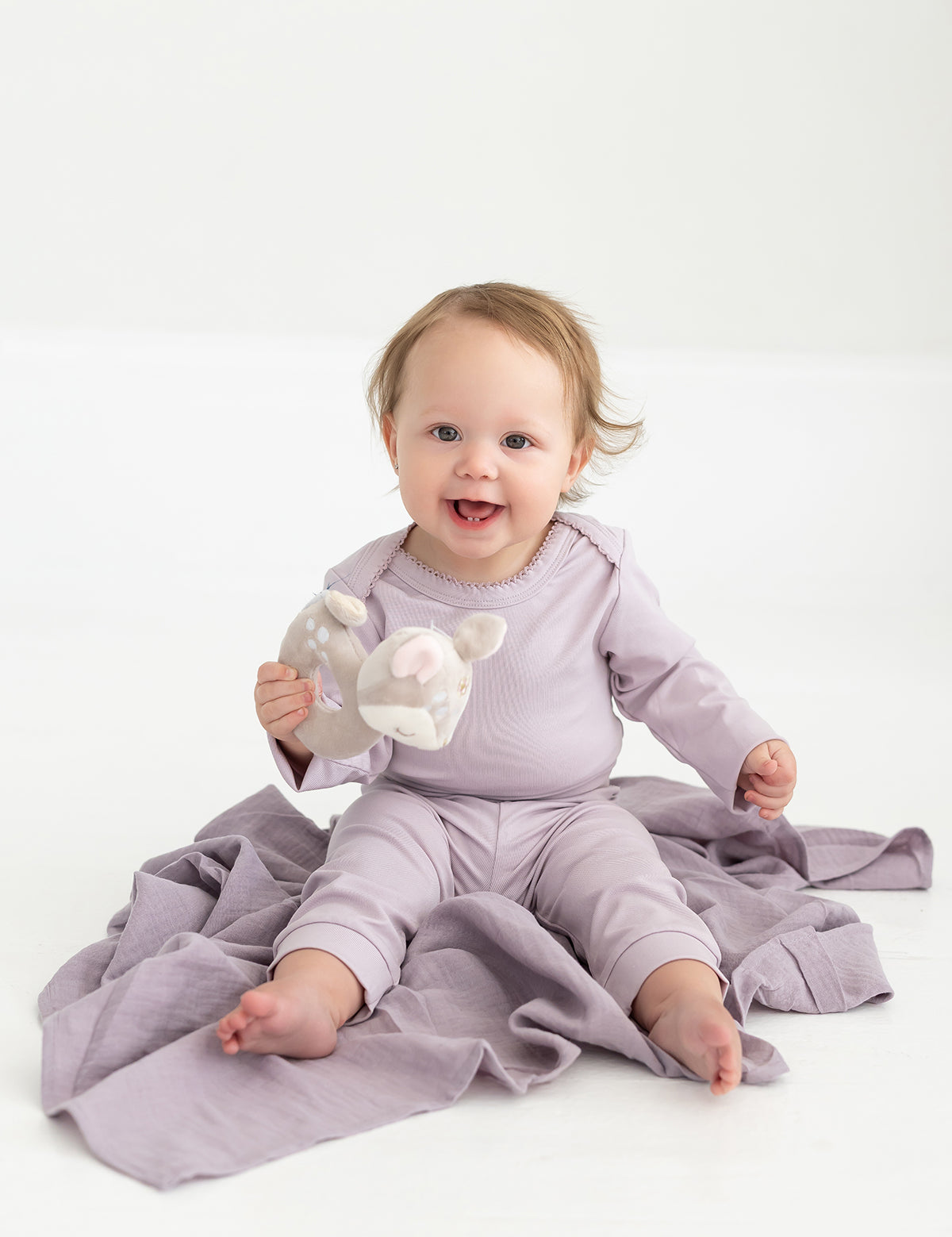 A smiling baby in a lavender outfit, featuring Mabel and Honey’s Soft Meadow Solid Ribbed Cuff Joggers with an elastic waistband made from a soft cotton blend, sits on a matching blanket and holds a stuffed animal toy against a plain white background.