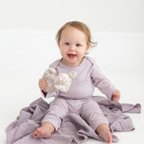 A smiling baby in a lavender outfit, featuring Mabel and Honey’s Soft Meadow Solid Ribbed Cuff Joggers with an elastic waistband made from a soft cotton blend, sits on a matching blanket and holds a stuffed animal toy against a plain white background.