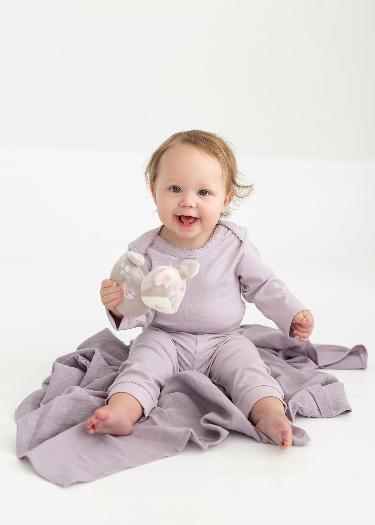 A smiling baby in a lavender outfit, featuring Mabel and Honey’s Soft Meadow Solid Ribbed Cuff Joggers with an elastic waistband made from a soft cotton blend, sits on a matching blanket and holds a stuffed animal toy against a plain white background.