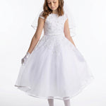 A young girl wearing the Lauren Marie Embroidered Bodice and Flower Beaded Waist Dress stands against a plain background, her gaze lowered as she gently holds the tulle skirt. The delicate lace bodice, paired with gloves and a soft veil, completes her graceful Communion look.