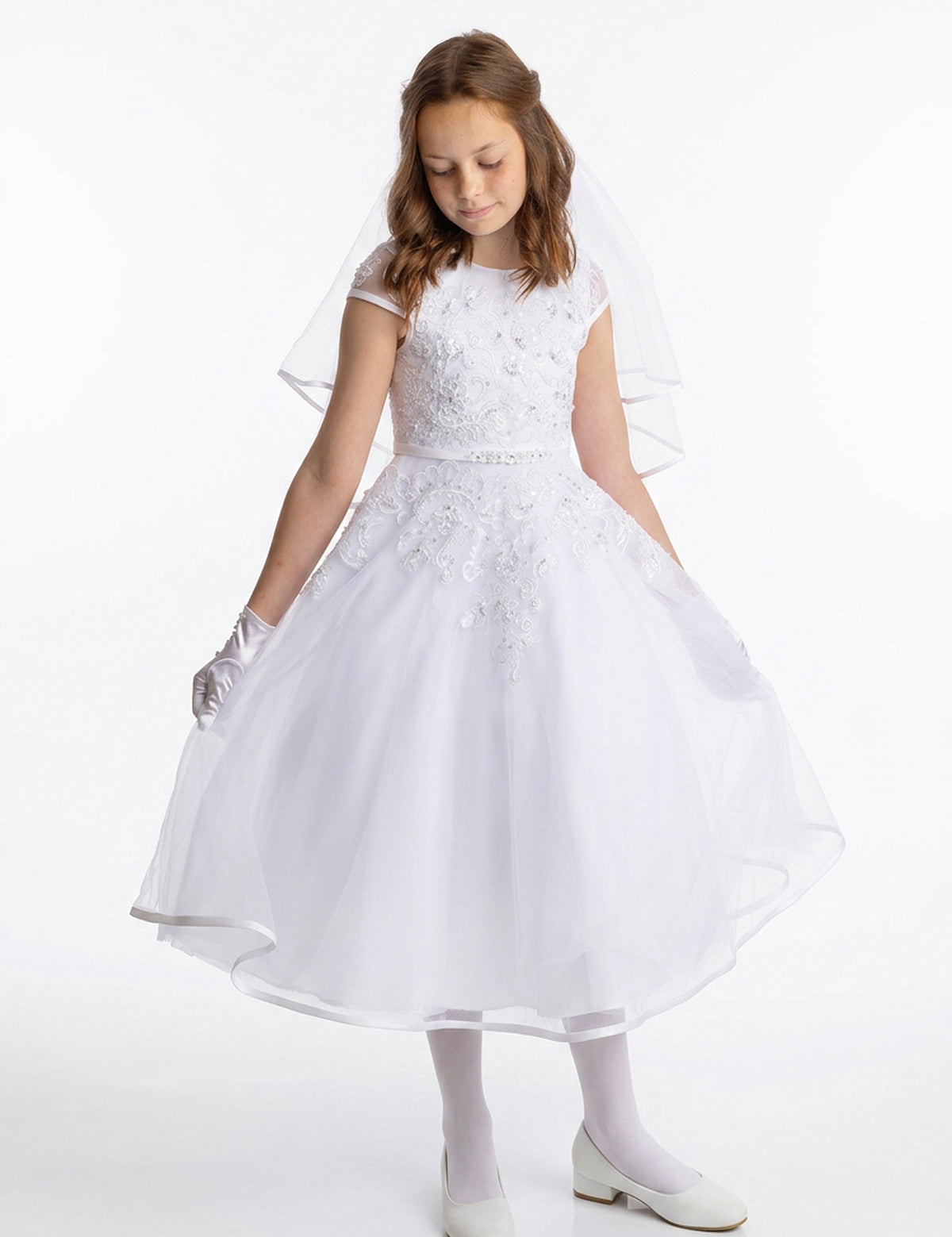A young girl wearing the Lauren Marie Embroidered Bodice and Flower Beaded Waist Dress stands against a plain background, her gaze lowered as she gently holds the tulle skirt. The delicate lace bodice, paired with gloves and a soft veil, completes her graceful Communion look.
