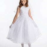 A young girl wearing the Lauren Marie Embroidered Bodice and Flower Beaded Waist Dress stands against a plain background, her gaze lowered as she gently holds the tulle skirt. The delicate lace bodice, paired with gloves and a soft veil, completes her graceful Communion look.
