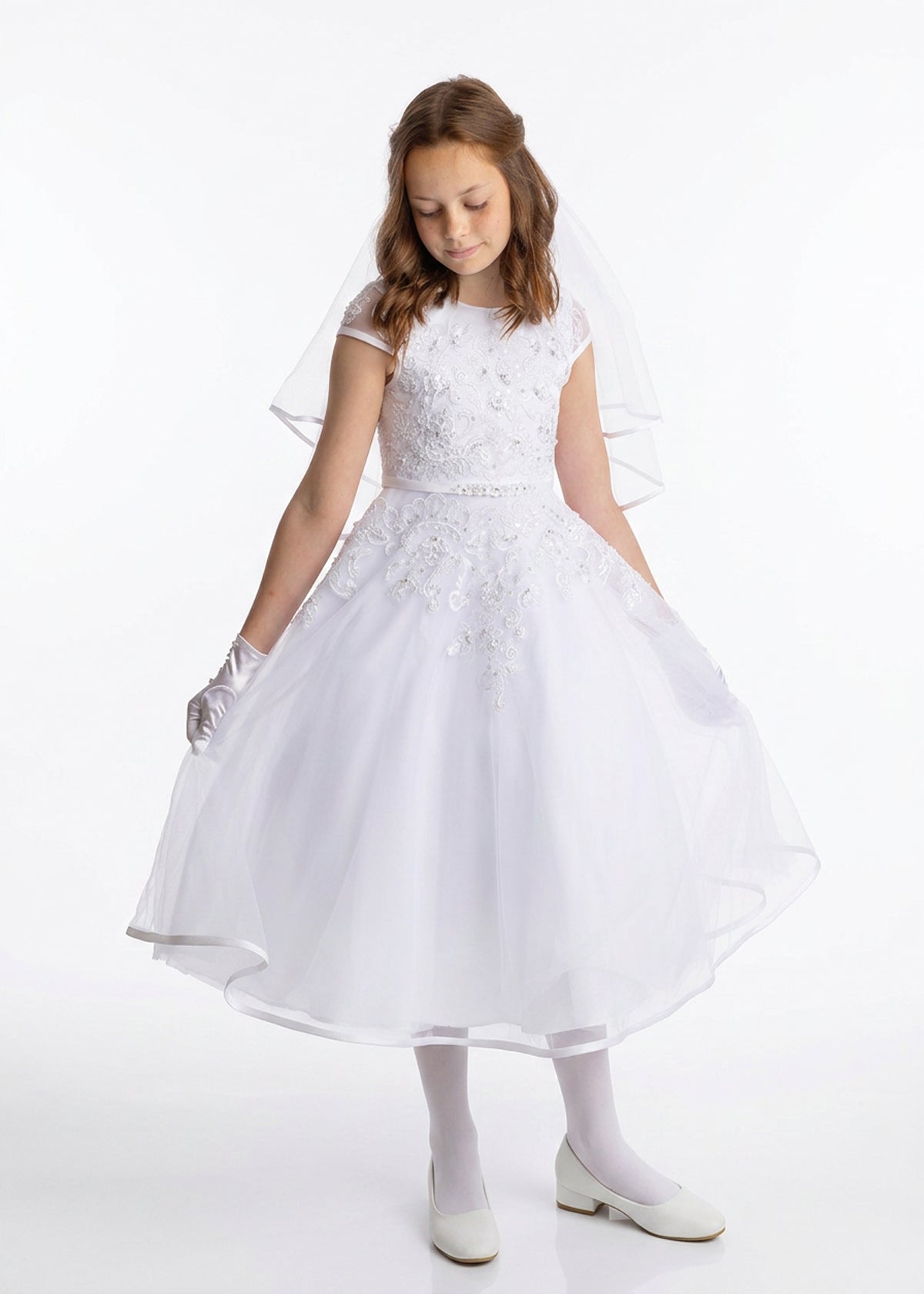 A young girl wearing the Lauren Marie Embroidered Bodice and Flower Beaded Waist Dress stands against a plain background, her gaze lowered as she gently holds the tulle skirt. The delicate lace bodice, paired with gloves and a soft veil, completes her graceful Communion look.