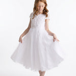 A young girl with long red hair wears the Lauren Marie “Scalloped Detail on Waist and Sleeve Dress,” a white, short-sleeved First Communion dress featuring a delicate lace bodice, scalloped detailing at the waist and sleeves, and an ankle-length organza tulle skirt. Paired with white shoes, she holds out the sides of her skirt and gazes down slightly against a plain white background.