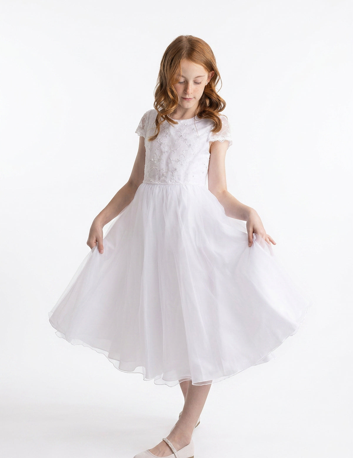 A young girl with long red hair wears the Lauren Marie “Scalloped Detail on Waist and Sleeve Dress,” a white, short-sleeved First Communion dress featuring a delicate lace bodice, scalloped detailing at the waist and sleeves, and an ankle-length organza tulle skirt. Paired with white shoes, she holds out the sides of her skirt and gazes down slightly against a plain white background.