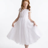 A young girl with long red hair wears the Lauren Marie “Scalloped Detail on Waist and Sleeve Dress,” a white, short-sleeved First Communion dress featuring a delicate lace bodice, scalloped detailing at the waist and sleeves, and an ankle-length organza tulle skirt. Paired with white shoes, she holds out the sides of her skirt and gazes down slightly against a plain white background.