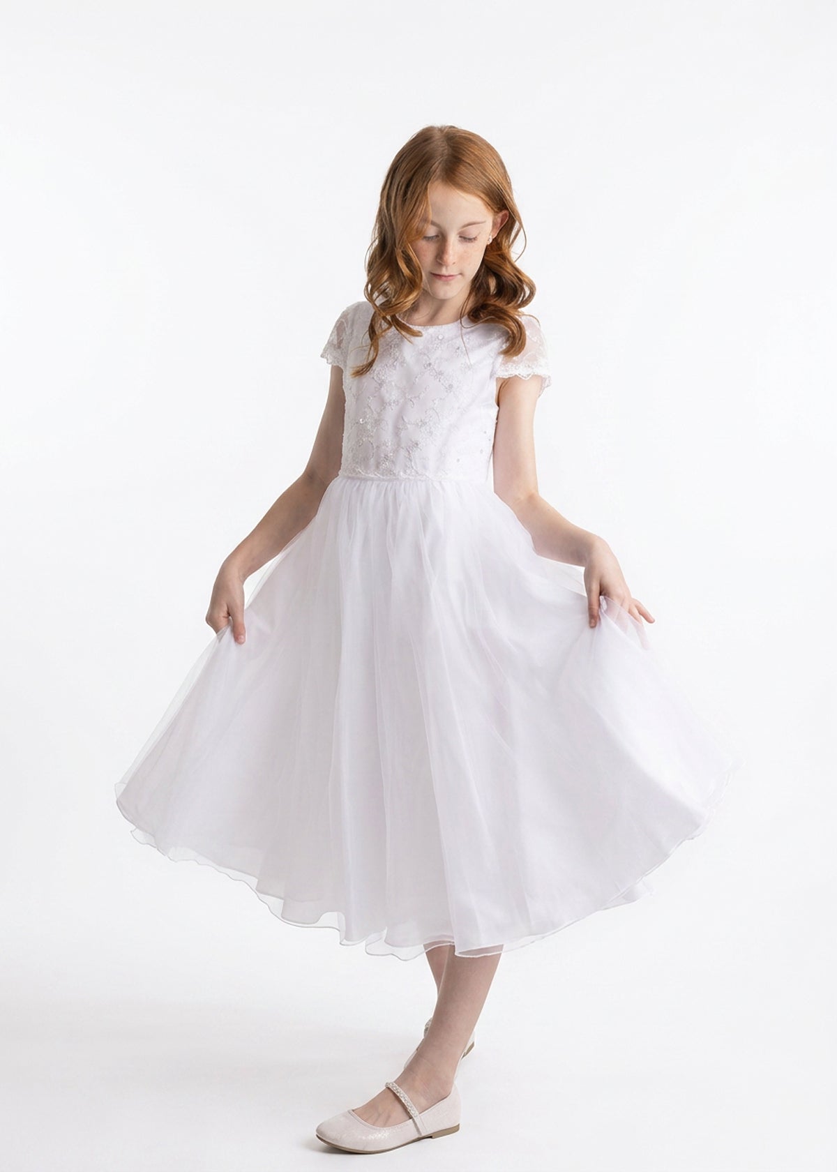 A young girl with long red hair wears the Lauren Marie “Scalloped Detail on Waist and Sleeve Dress,” a white, short-sleeved First Communion dress featuring a delicate lace bodice, scalloped detailing at the waist and sleeves, and an ankle-length organza tulle skirt. Paired with white shoes, she holds out the sides of her skirt and gazes down slightly against a plain white background.