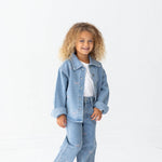 A young child with curly hair smiles while wearing a light blue denim jacket and the Mabel + Honey Bow Embroidered Rhinestone Wide Leg Denim Jeans (Big Kid), featuring a trendy wide leg silhouette. They pair the look with a white shirt and white sneakers, standing against a plain white background.