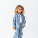 A young child with curly hair smiles while wearing a light blue denim jacket and the Mabel + Honey Bow Embroidered Rhinestone Wide Leg Denim Jeans (Big Kid), featuring a trendy wide leg silhouette. They pair the look with a white shirt and white sneakers, standing against a plain white background.