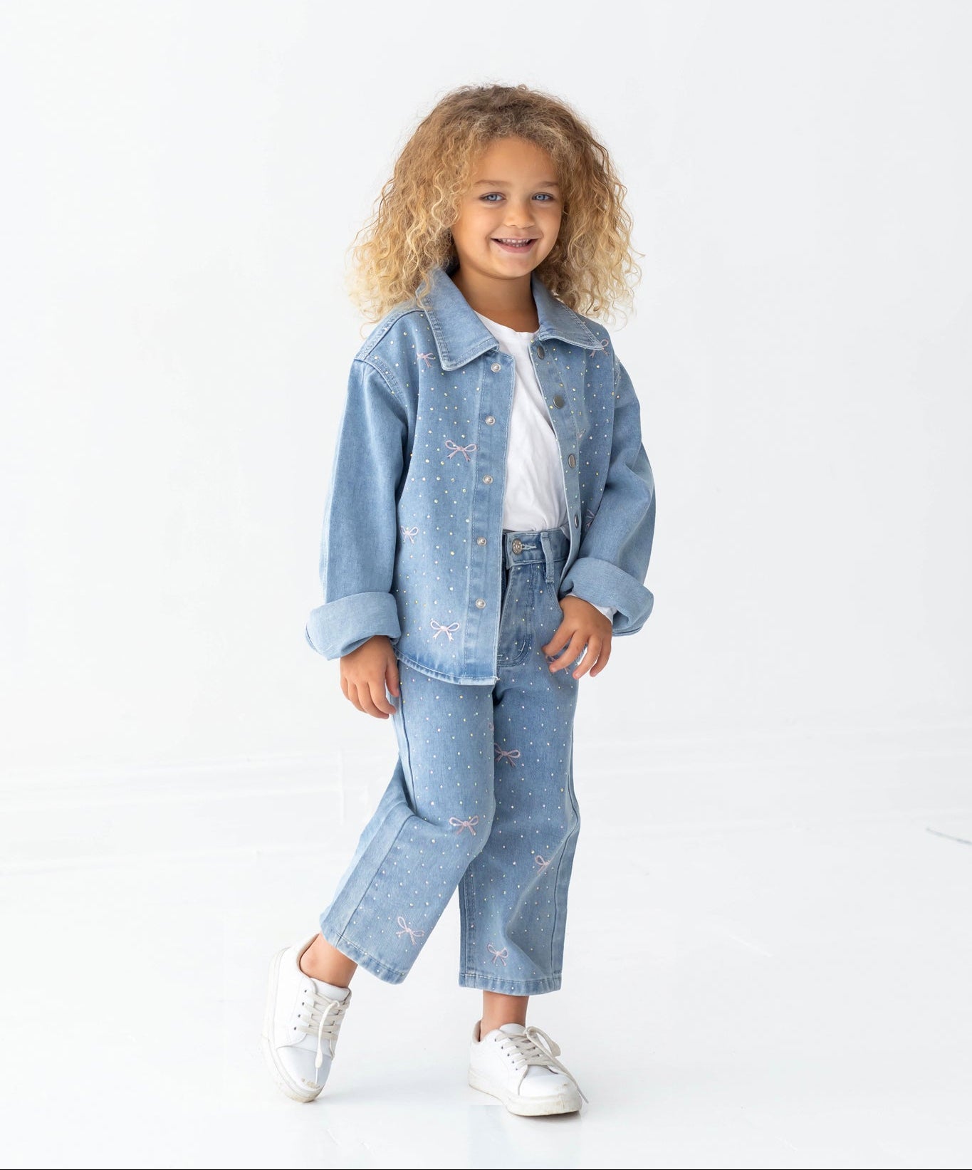 A smiling young child with long, curly blonde hair poses against a white background, wearing Mabel and Honey’s Bow Embroidered Rhinestone Wide Leg Denim Jeans (Big Kid), featuring small pearl-like studs and pink bow accents. The look is completed with a light blue denim jacket, a white t-shirt, and white sneakers.