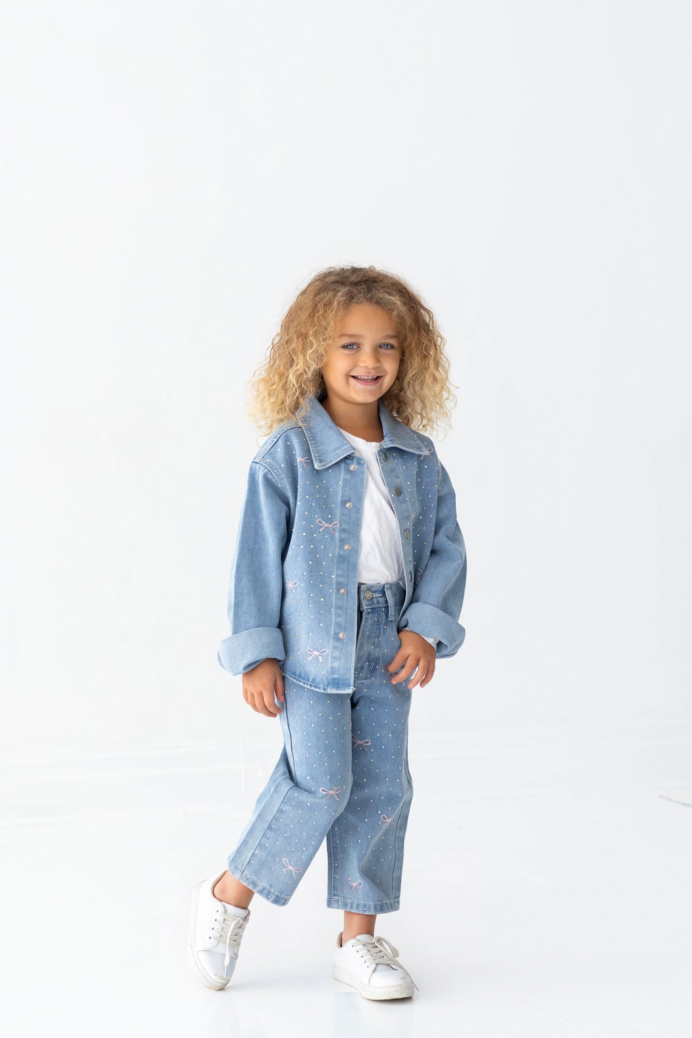A young child with curly hair smiles while wearing a light blue denim jacket and the Mabel + Honey Bow Embroidered Rhinestone Wide Leg Denim Jeans (Big Kid), featuring a trendy wide leg silhouette. They pair the look with a white shirt and white sneakers, standing against a plain white background.