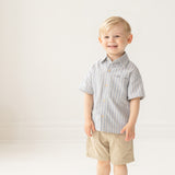 A young boy with blonde hair smiles in a bright room, wearing the Breezy Blue Skies Top by Mabel and Honey— a light blue and white striped shirt— paired with khaki shorts and gray shoes against a plain white background.