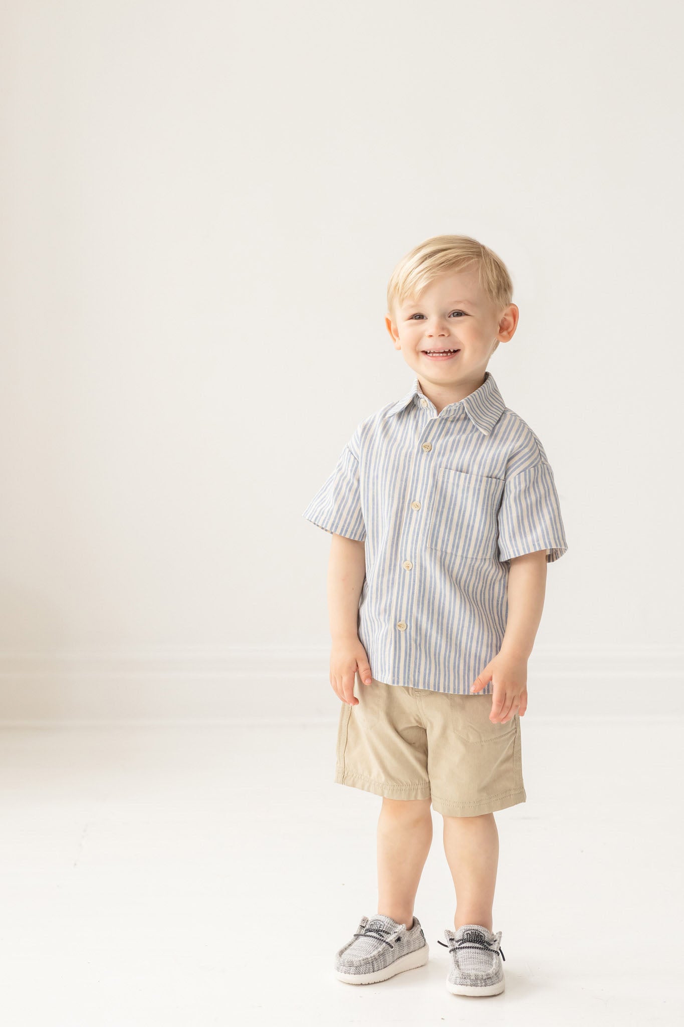 A young boy with blonde hair smiles in a bright room, wearing the Breezy Blue Skies Top by Mabel and Honey— a light blue and white striped shirt— paired with khaki shorts and gray shoes against a plain white background.